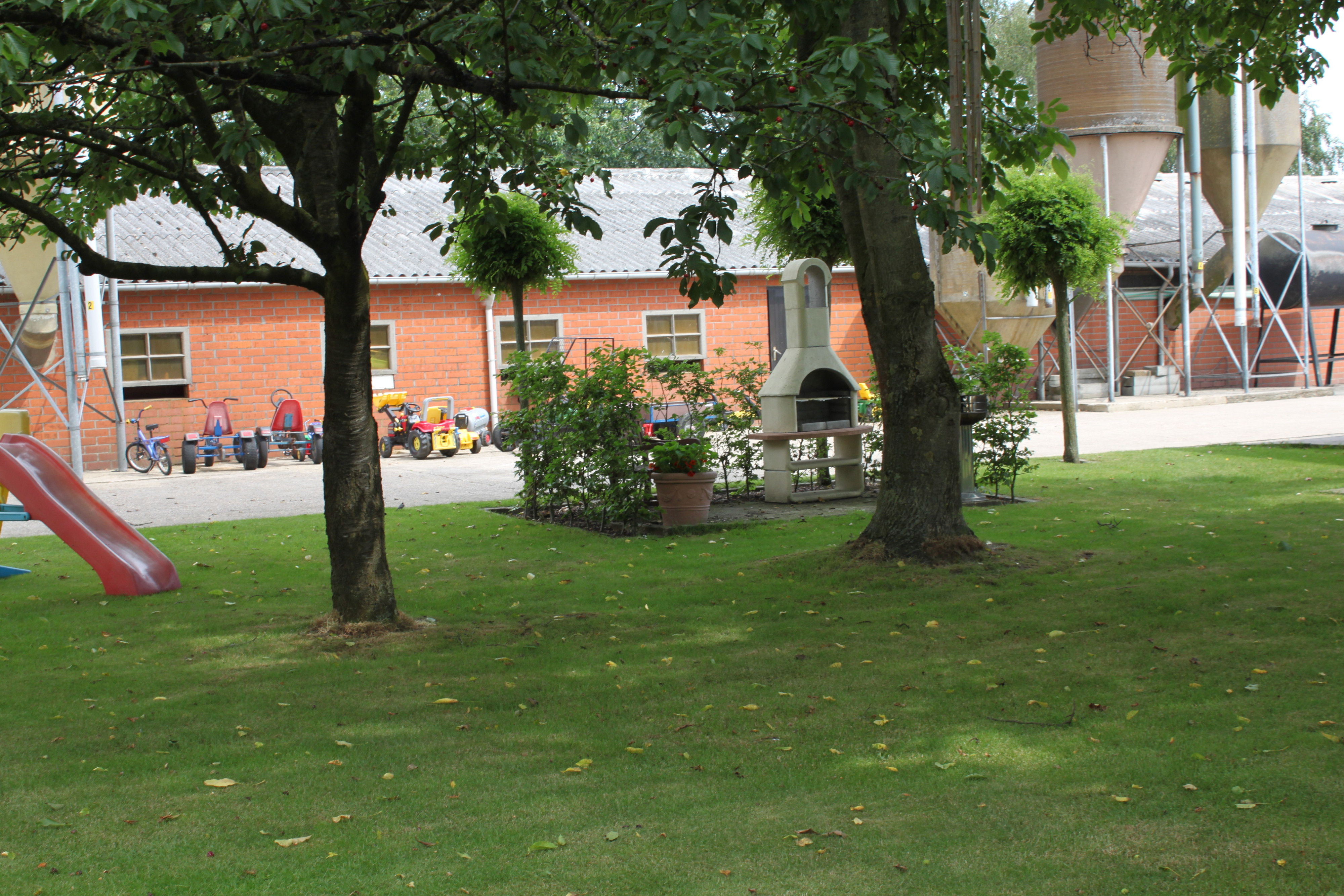 foto van grasplein met twee bomen, glijbaan, barbecue. Op de achtergrond staat een gebouw en silo's