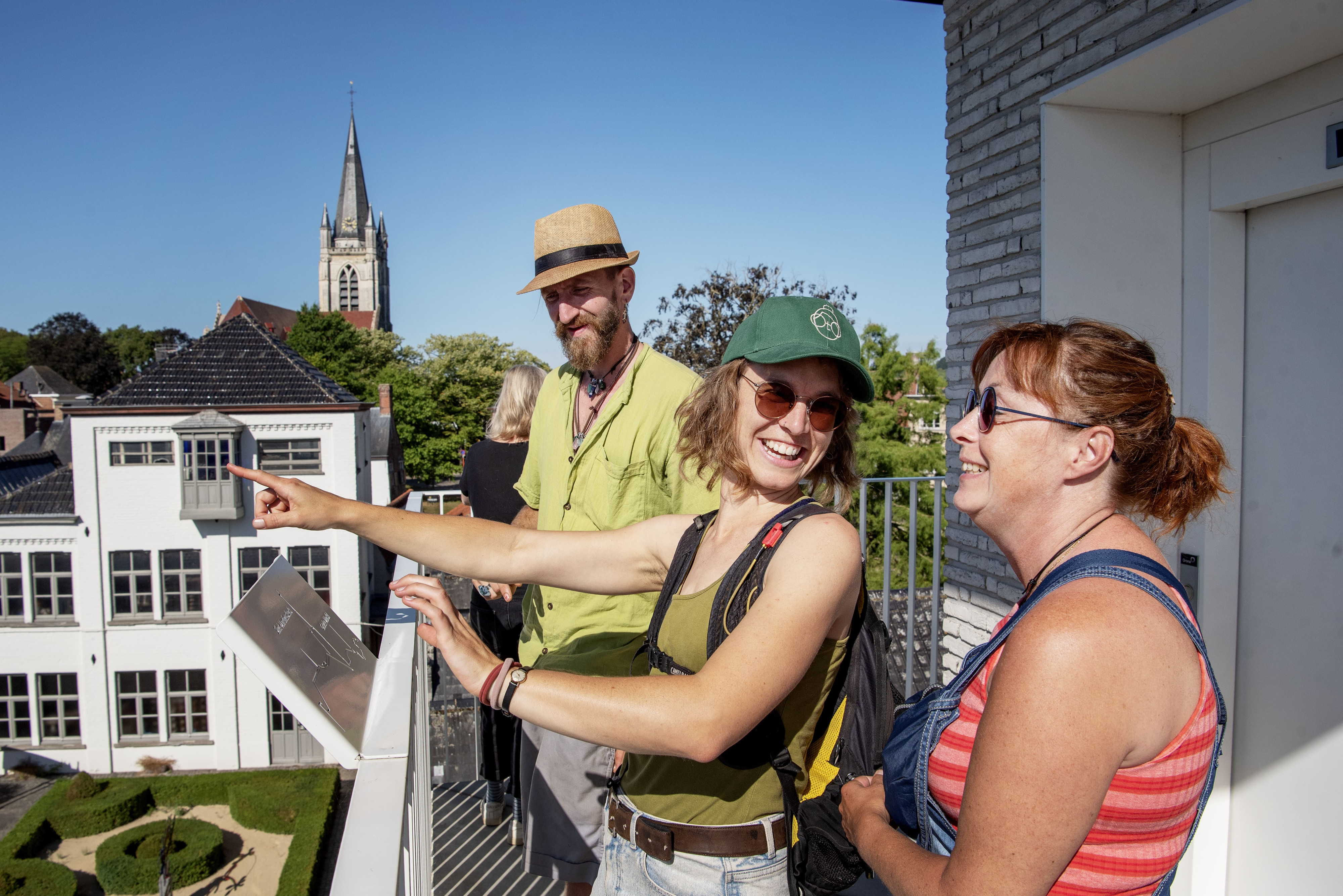 3 mensen staan op balkon, middelste vrouw wijst naar iets en ze lachen alle drie