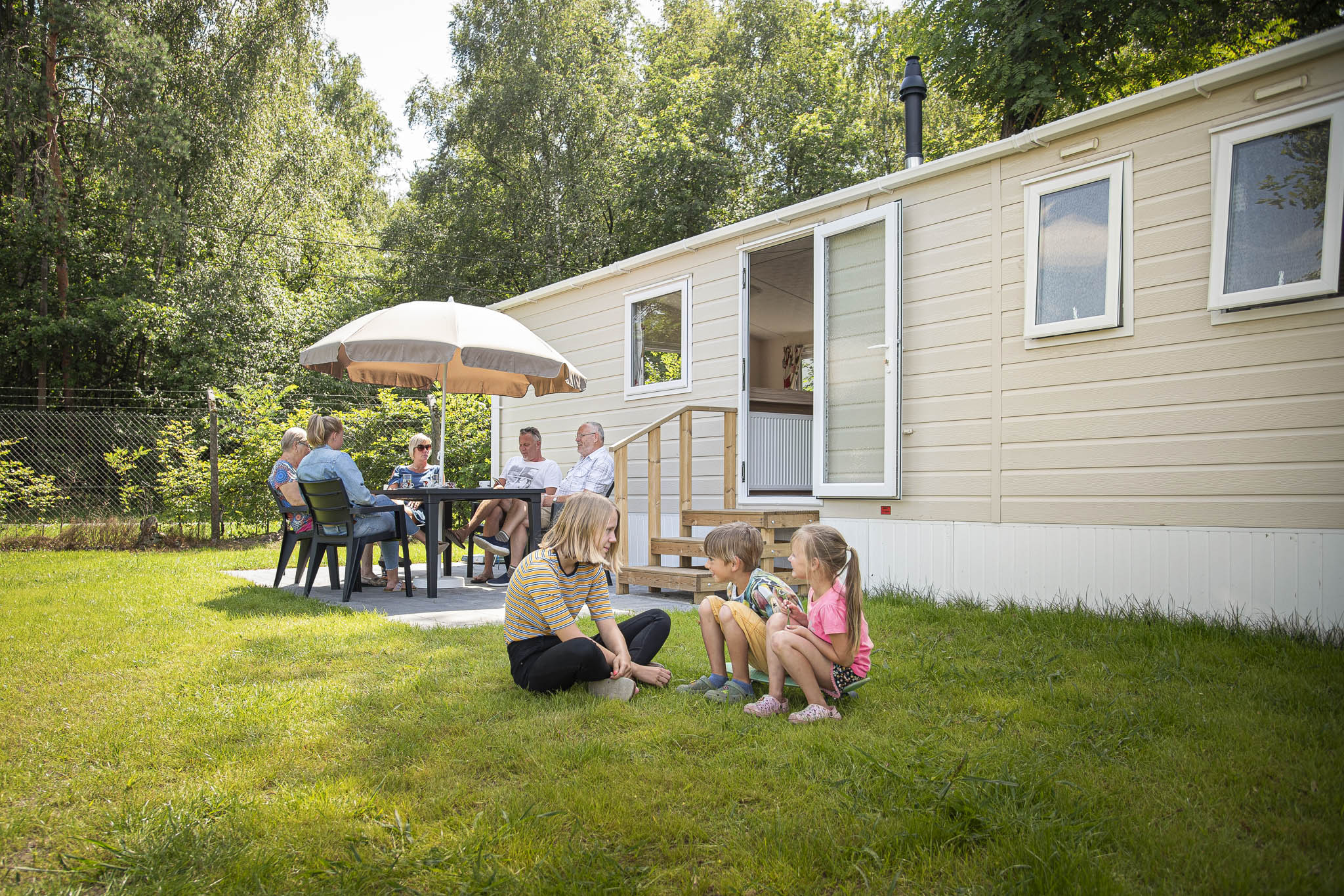 foto van drie kinderen voor stacaravan. Op de achtergrond zitten 4 volwassenen aan tafel onder parasol