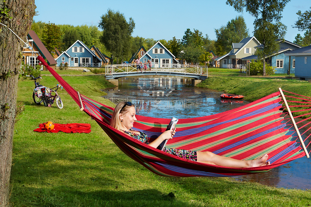 dame zit te lezen in hangmat, we zien een grasveld, beek, gebouwen en bossen op de achtergrond